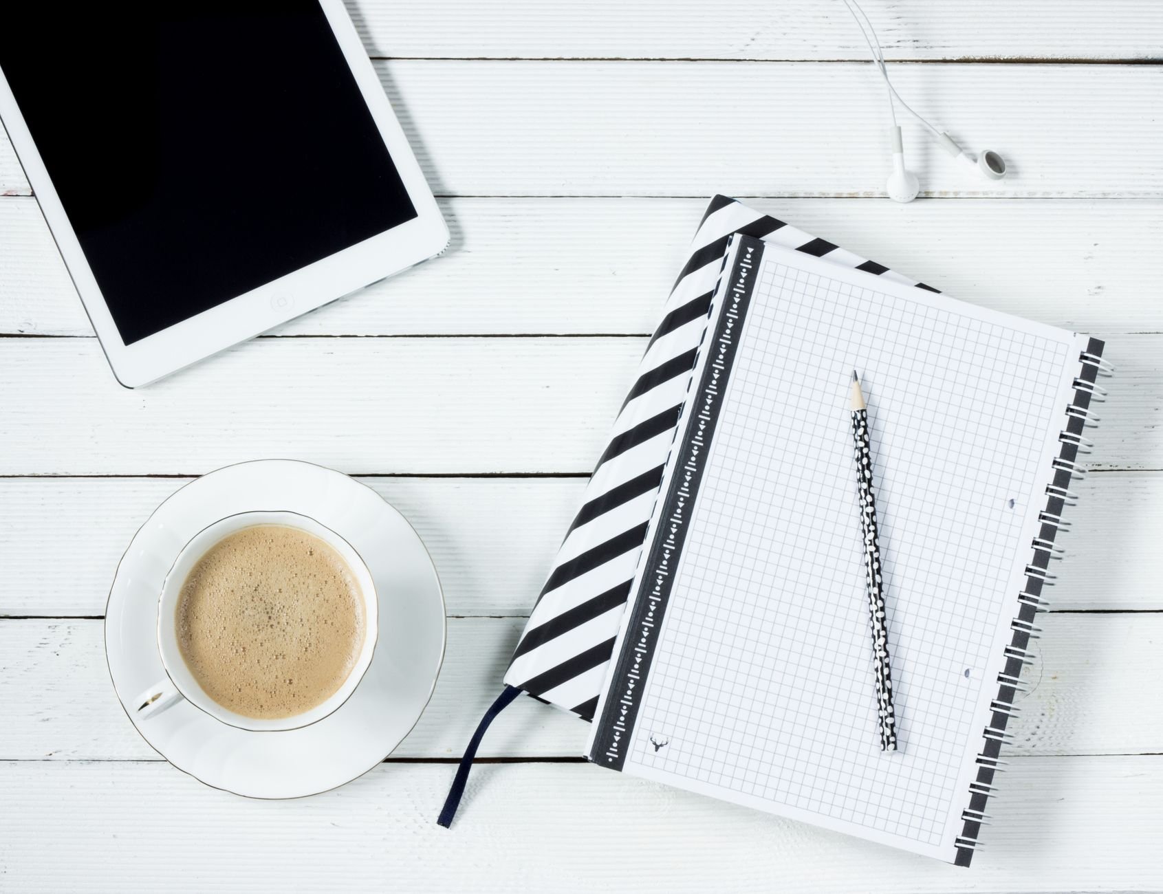 Flatlay of a modern workspace featuring coffee, tablet, and notebook with pencil on a white wooden desk.